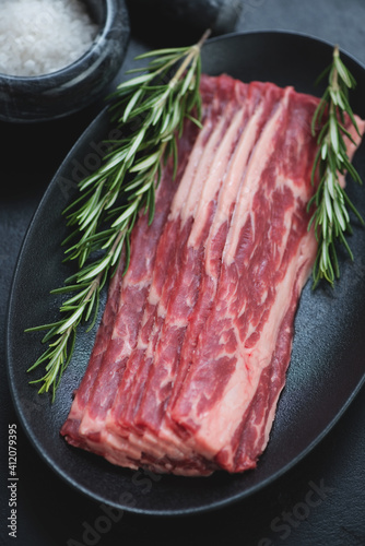 Close-up of raw fresh bacon made of marbled beef with rosemary on a black plate, vertical shot, selective focus