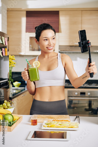 Smiling pretty fitness blogger filming herself drinking mug of green smoothie she made at home photo