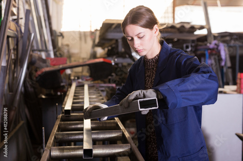Confident young woman in blue overalls working in mechanical metalworking workshop, checking metal structures..