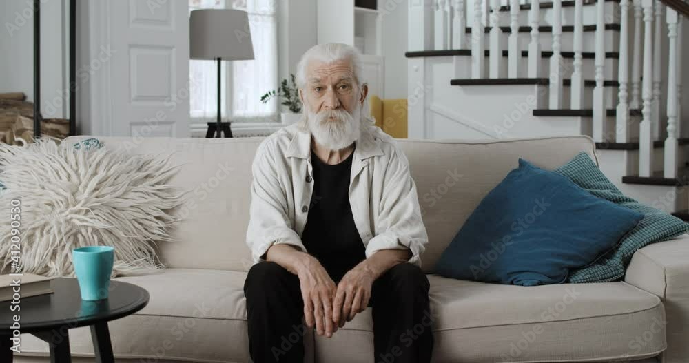 Crop view of elderly grey haired man in 70s turning head and looking to camera. Portrait of bearded grandfather posing while sitting on sofa at stylish home. Concept of retirement