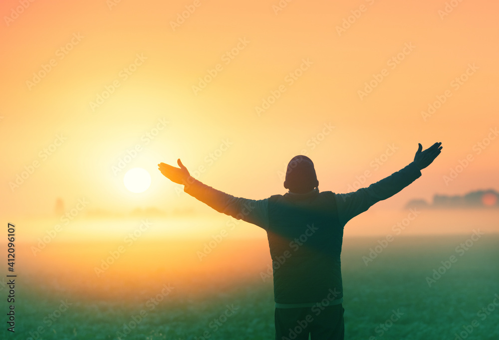 A man with raised arms stands in a field in the early morning and looks at the sunrise
