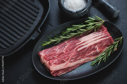 Raw marbled beef bacon with fresh rosemary and salt, studio shot over black stone background with a cast-iron grill