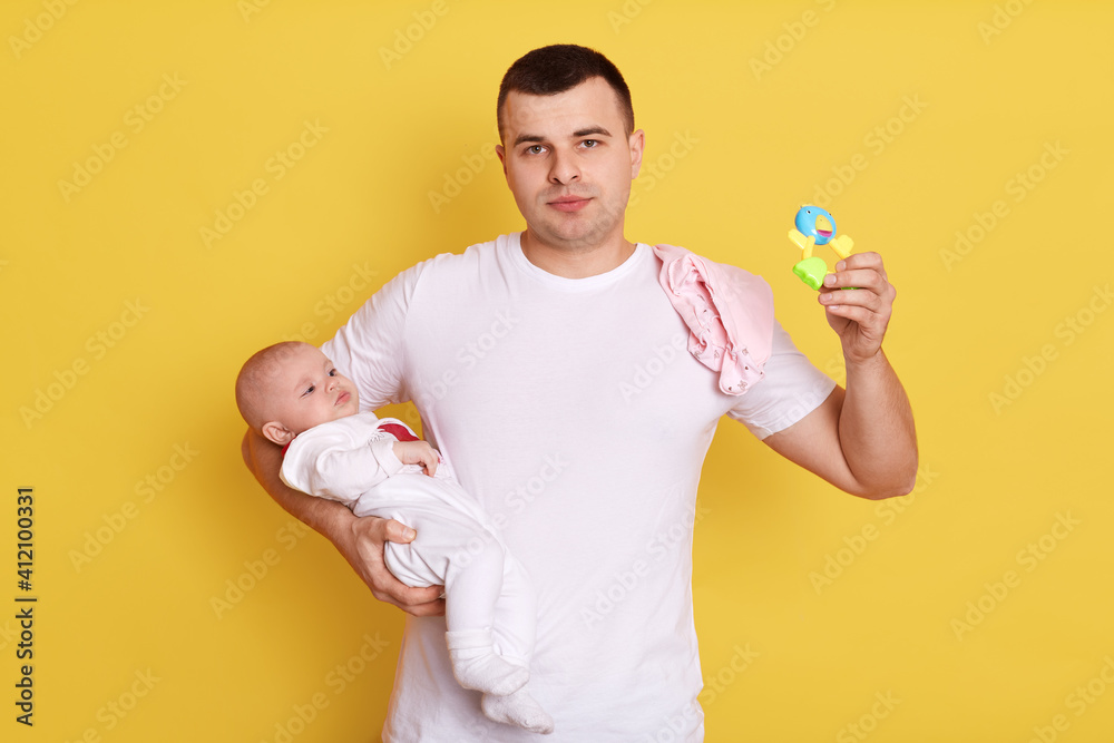 Handsome father and baby child posing isolated over yellow background, dad holds beanbag in hands, wearing casual white t shirt, european daddy newborn daughter. Handsome father and baby child posing isolated over yellow background, dad holds beanbag in hands, wearing casual white t shirt, european daddy newborn daughter.