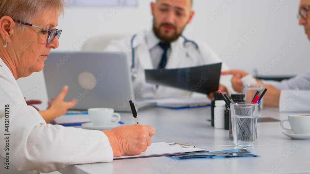 Close up of senior woman doctor taking notes on clipboard while radiologist coworkers discussing in background analysing x-ray and writing on laptop. Profesional teamworker having medical meeting Close up of senior woman doctor taking notes on clipboard while radiologist coworkers discussing in background analysing x-ray and writing on laptop. Profesional teamworker having medical meeting