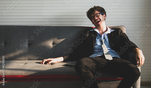 handsome caucasian businessman wearing black suit with loosen necktie sitting on a couch leaning against a backrest laughing happily. Success, employment concept
