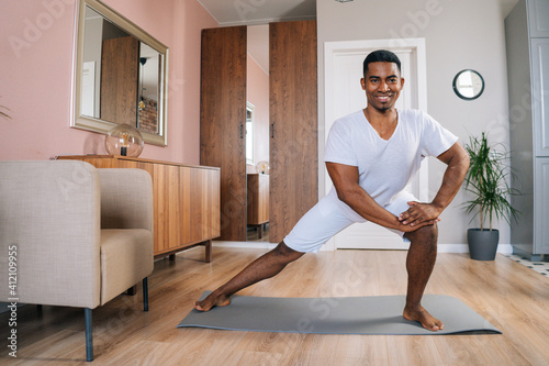Front view of smiling strong African-American man doing side lunge exercise at home during working out standing on yoga mat at domestic room, looking at camera. Concept of sport training at home gym.