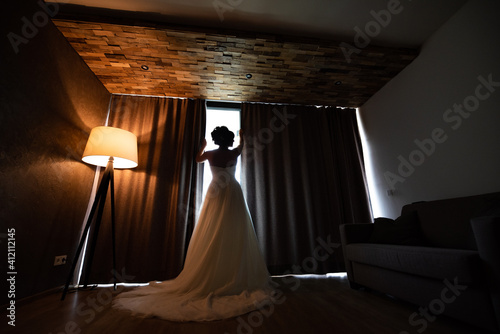 The bride in a wedding dress with her back turned, looking out the window. The interior is dark next to the lit lamp and she is wearing a white long wedding dress.