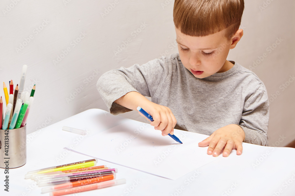 Cute little boy with blond hair draws colored pencils at home. Draws at the white table. Cute little boy with blond hair draws colored pencils at home. Draws at the white table.