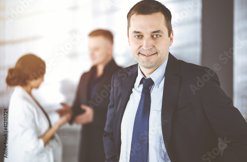 Portrait of a self-confident middle aged businessman in a blue suit, standing in a sunny modern office with his colleagues at the background. Concept of business success