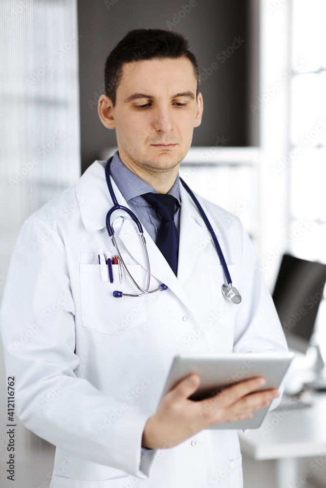 Friendly male doctor using tablet computer in clinic near his working place. Perfect medical service in hospital. Medicine and healthcare concept Friendly male doctor using tablet computer in clinic near his working place. Perfect medical service in hospital. Medicine and healthcare concept