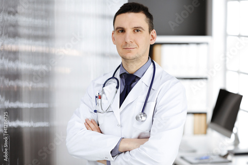 Male doctor standing straight with arms crossed in clinic near his working place. Perfect medical service in hospital. Medicine and healthcare concept