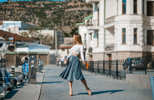 An attractive young fashionable woman dressed in a stylish spring-summer outfit walks along the embankment among the yachts at a luxury resort. Portrait of a beautiful cute girl on a sunny day. photo