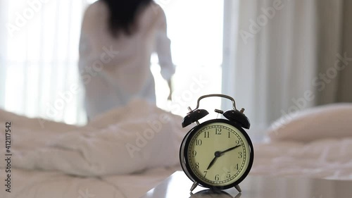 Closeup alarm clock having a good day with background happy woman stretching in bed after waking up, sunlight in morning.