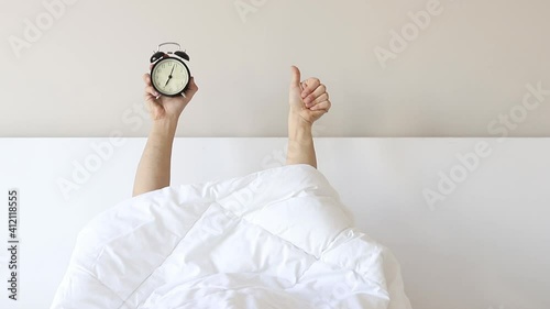 Man showing arm raised up black alarm clock behind duvet in the bed room, Young boy with two hands sticking out from the blanket. wake up with fun in morning concept.