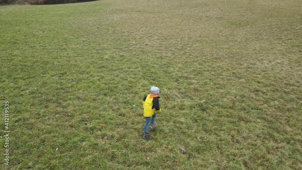 Boy with yellow jacket is running through meadow and having fun and a drone is following and filming him doing it Boy with yellow jacket is running through meadow and having fun and a drone is following and filming him doing it