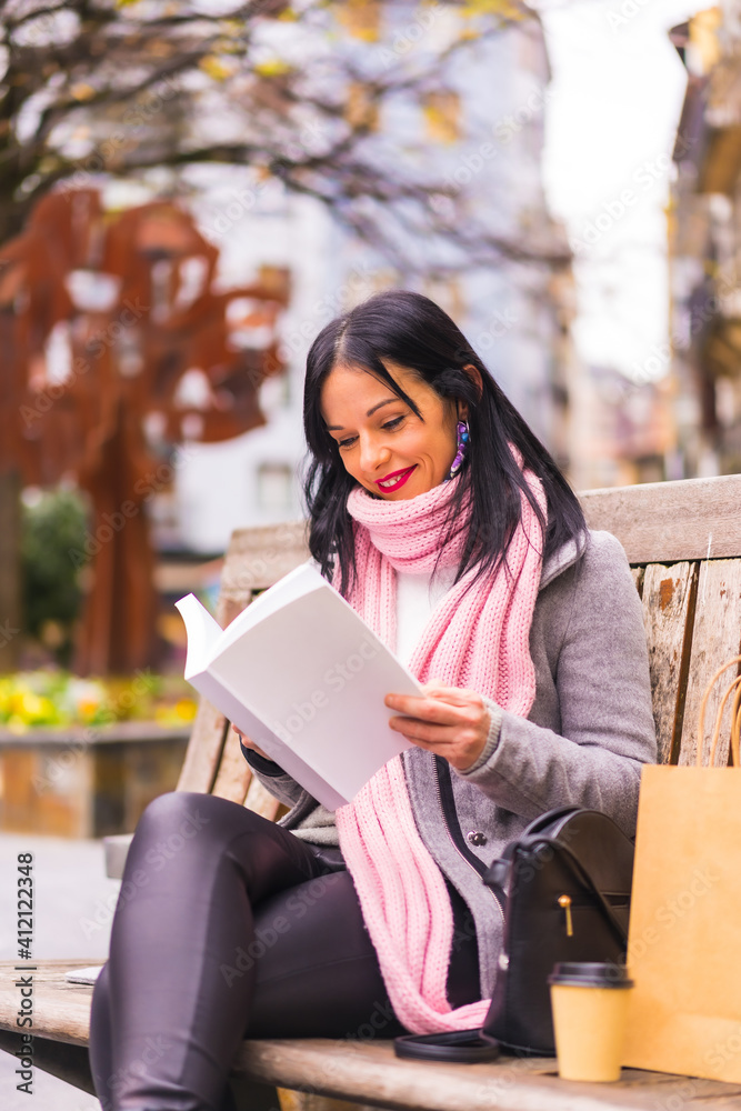 Fototapeta premium Lifestyle, Caucasian brunette girl reading a book in a park, smiling sitting on a bench