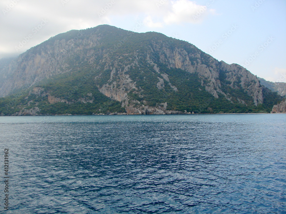 Landscape of the calm azure surface of the sea bay surrounded by mountain forests under a cloudy morning sky.