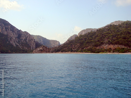Landscape of the calm azure surface of the sea bay surrounded by mountain forests under a cloudy morning sky.