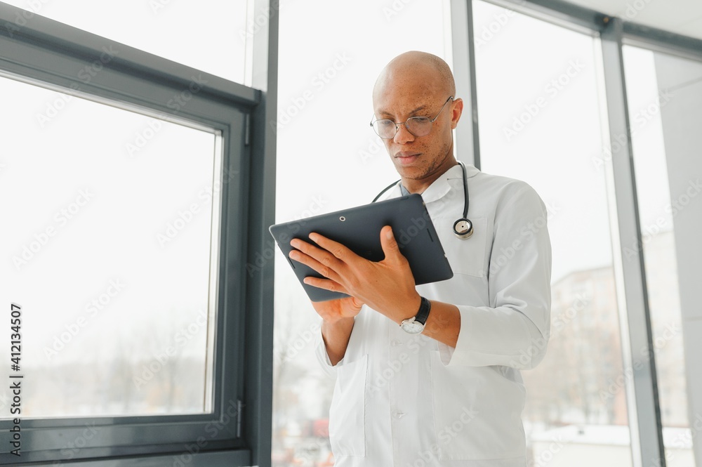 Mature african doctor using digital tablet in corridor . Portrait of confident male doctor using tablet computer in clinic with copy space. Successful smiling doctor in labcoat wearing stethoscope.