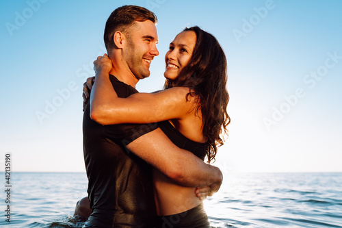 Happy young fit couple in the sea or ocean hug each other with love at summer sunset. Romantic mood, tenderness, relationship, vacation concept.