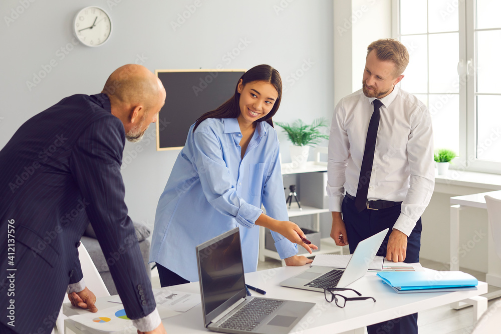 Cheerful girl points to a laptop screen to her excited colleagues in a bright modern office.