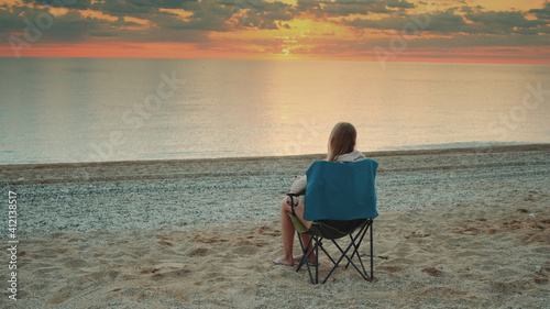 Woman sitting on camping chair and drinking coffee. Relaxing and enjoying nature. Admiring sunrise on the sea.