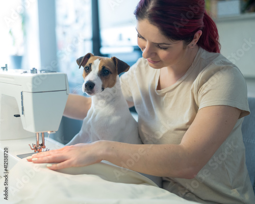 Caucasian woman sews while sitting in the kitchen. Dog Jack Russell Terrier sits on the lap of the owner. Home hobby.