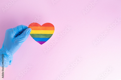 A hand in a protective blue glove holds a rainbow paper heart on a pink background. Safe holiday greetings concept. Safe Valentine's Day 2021 concept. LGBTQL concept photo