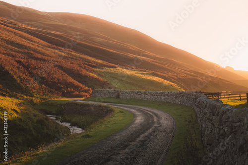Golden sunset or sunrise light on an empty country road alongside an old stone wall on the Glencorse walk in the Pentland Hills Regional Park countryside in Edinburgh, Midlothian, Scotland. photo