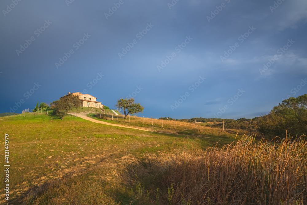 A lone rural countryside farmhouse in Radicondoli catches some golden sunset or sunrise light with a moody dark blue sky as a backdrop in Tuscany, Italy.