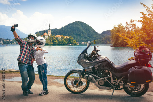 Couple in love travel together takes a selfie on a smartphone. Stands with motorcycle with bags. Tourism and vacation. Sunny day. Bled lake, island castle and mountains in background, Slovenia Europe