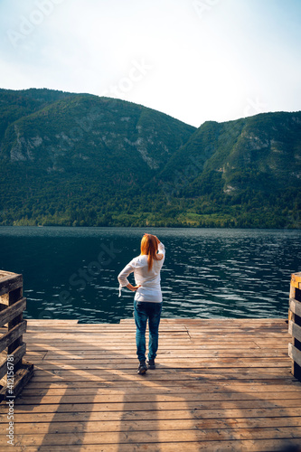 The female, girl, woman stand on wooden pier. Mountain lake landscape, Green forest, sunset time. travel and vacation, vertical photo