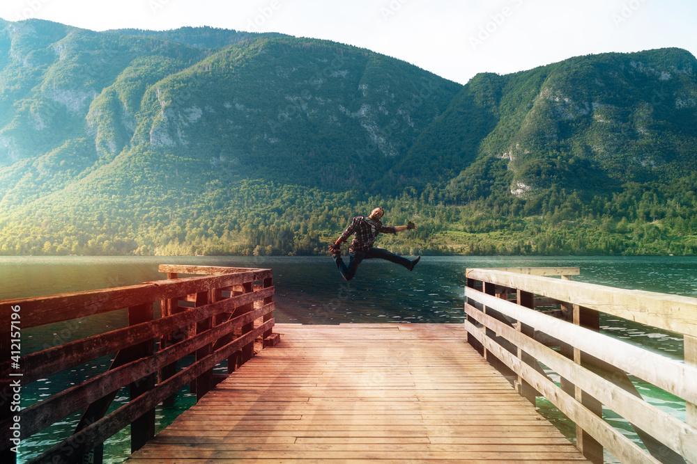 Man happy jump alone standing on a wooden pier at a mountains over water. Summer sunset time. Travel and freedom concept 