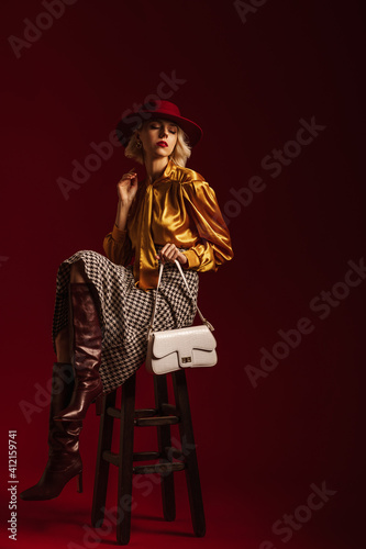 Fashionable woman wearing hat  yellow blouse  houndstooth print skirt  leather high heeled boots  holding small stylish white bag  posing on dark red background