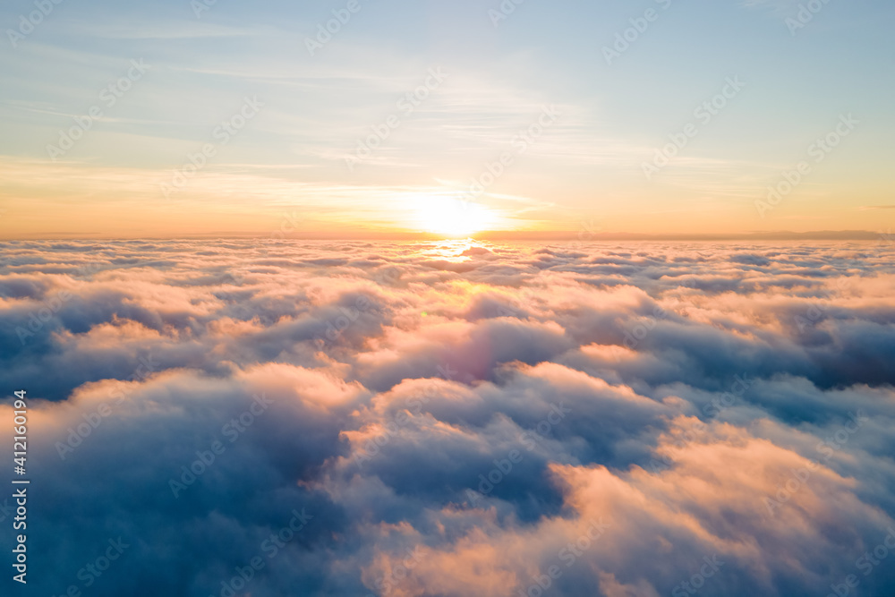 Aerial view of bright yellow sunset over white dense clouds with blue sky overhead.