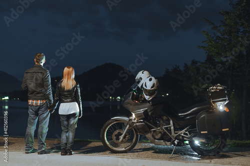 Motorcyclists couple in love travel together. Stands by a tourer motorcycle with bags. Tourism and vacation. Summer night. Bled lake, island and mountains in background, back view. Slovenia, Europe