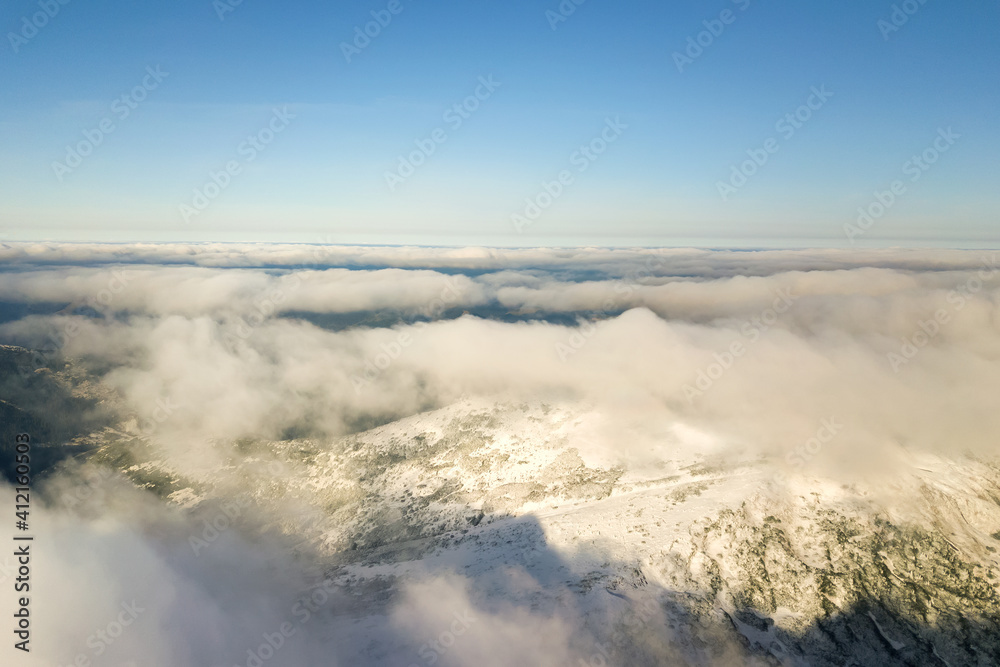 Obraz premium Aerial view from above of white puffy clouds covering snowy mountain tops in bright sunny day.