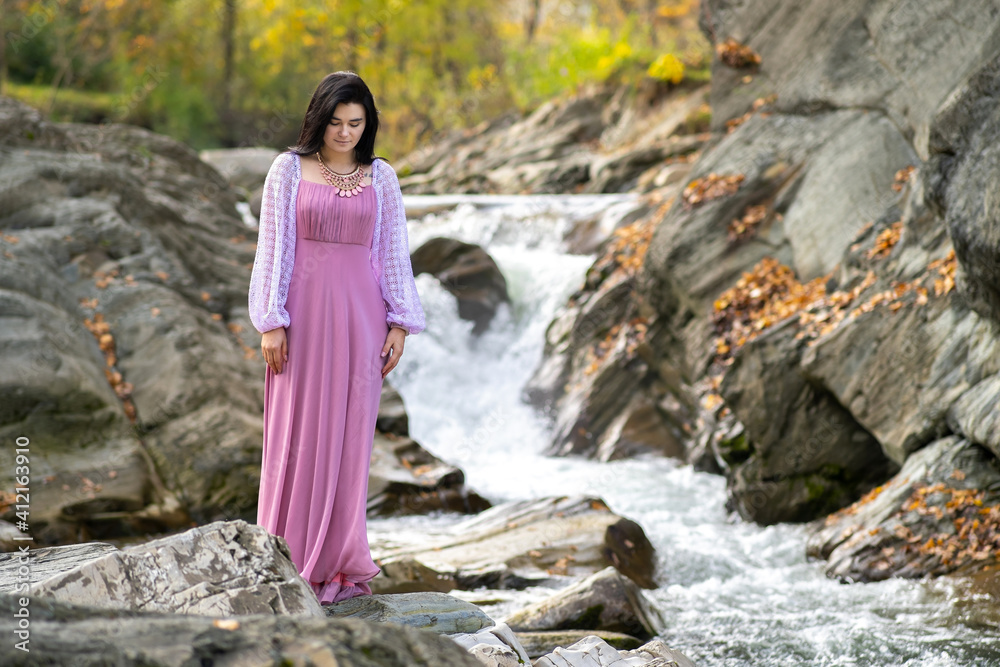 Young pretty woman in long fashionable dress standing near small mountain river with fast moving water. Young pretty woman in long fashionable dress standing near small mountain river with fast moving water.
