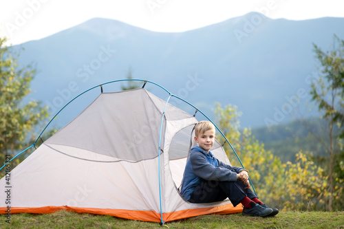 Happy child boy resting in a tourist tent at mountain campsite enjoying view of beautiful summer nature. Hiking and active way of life concept.