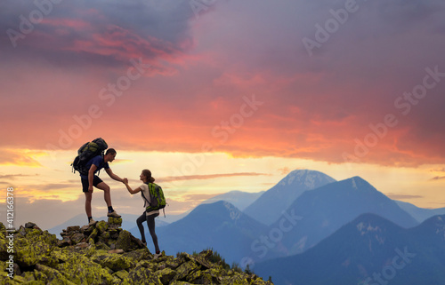 Tourist couple helping each other to climb high rock in evening mountains at sunset.