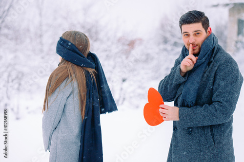 Handsome elegant man, finger to mouth - tsss gesture, surprise for his girlfriend holding red heart, greeting card with space. Couple in love on a walk in a winter park, valentine's day or celebration photo