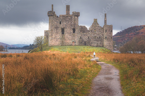 A distant young female tourist approaches the historic ruins of Kilchurn Castle  at Loch Awe in Argyll and Bute on a moody day in the Scottish Highlands  Scotland.