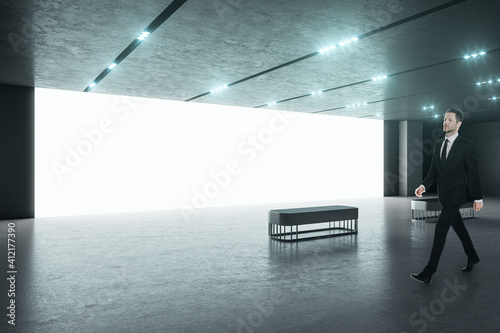 Businessman in black suit in a spacious modern hall with big blank white screen wall, concerete flloor and benches. Business presentation concept. Mockup photo