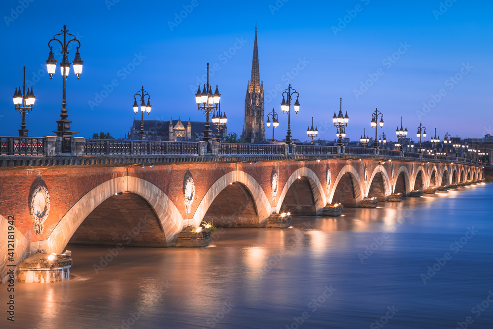 The historic Pont de Perre illuminated at night over the River Gardonne in Bordeaux, France with Bordeaux Cathedral in the background.