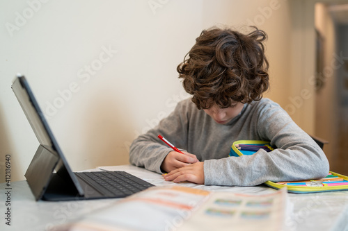 A schoolboy is studying o the kitchen table watching the lessons remotely online and writing on the paper book. Elementary remote school’s learning concept. photo