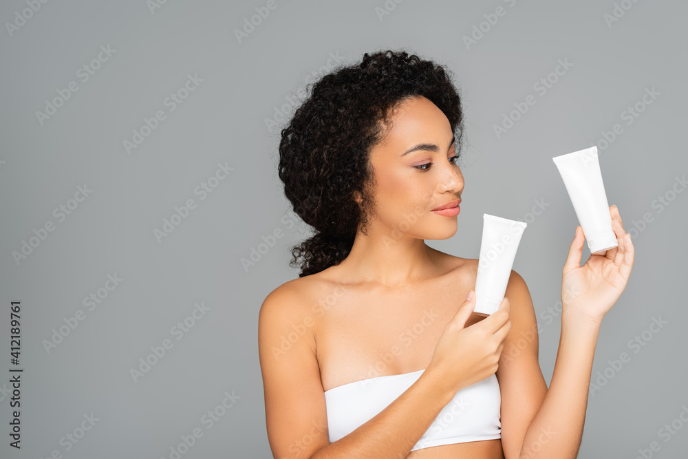 African american woman in white top holding tubes with cosmetic cream isolated on grey African american woman in white top holding tubes with cosmetic cream isolated on grey