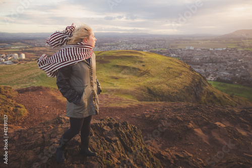 Photo A young blonde female tourist with a scarf looks out to a cityscape view of Edinburgh, Scotland on a windy afternoon up Arthur's Seat