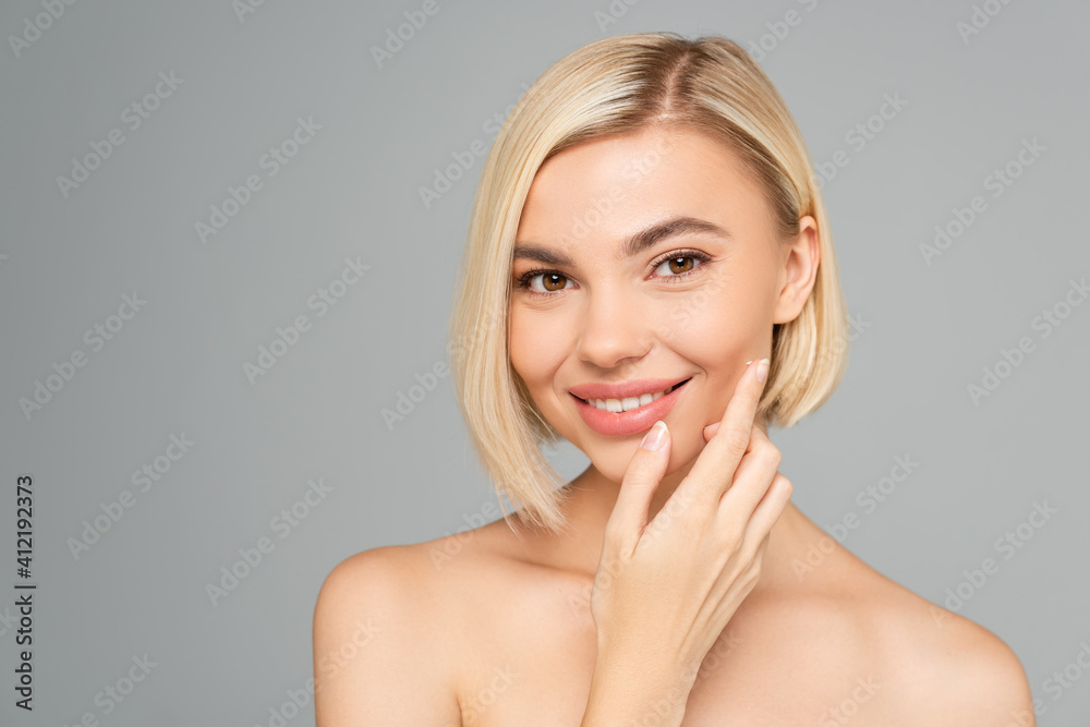 Smiling blonde woman applying cosmetic cream on cheek isolated on grey Smiling blonde woman applying cosmetic cream on cheek isolated on grey