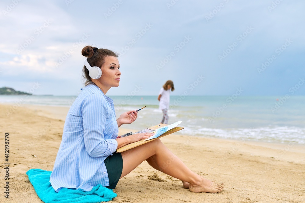 Mature woman sitting on sandy beach drawing sketch of sea