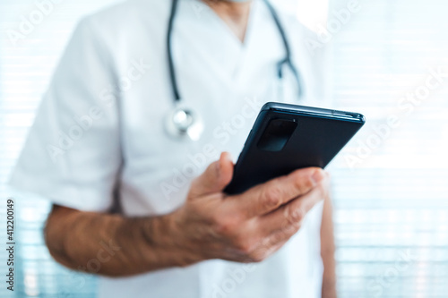 Mature male doctor - nurse, looking at mobile phone next to a hospital window. Covid-19 and medicine concept